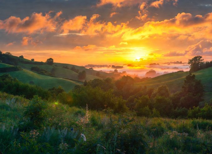 A picturesque landscape at sunset with vibrant orange and yellow clouds over rolling green hills, scattered trees, and a misty valley.