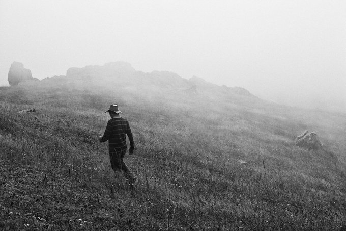 A black and white photograph showing a person wearing a wide-brimmed hat and a striped shirt walking through tall grass on a misty hillside with rocky outcroppings.