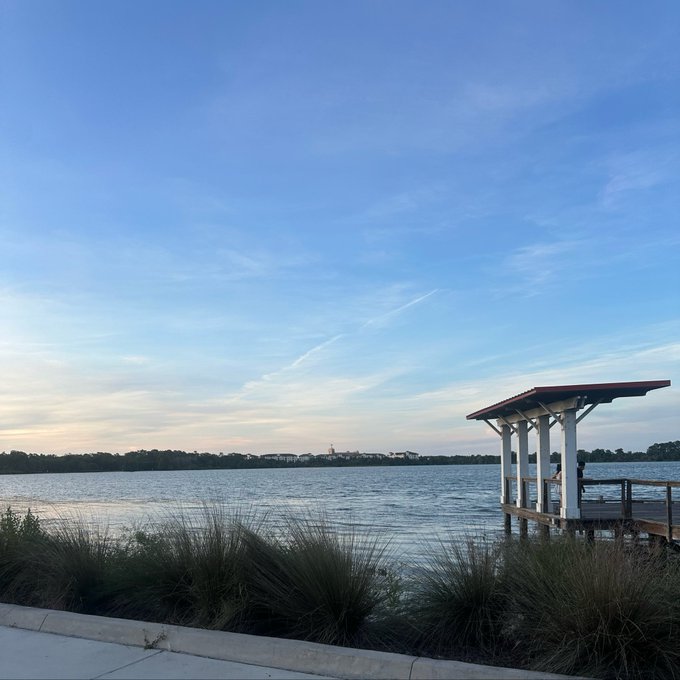 The image features a serene lakeside scene with a pier extending into calm waters, surrounded by lush green grass and bushes. A person can be seen sitting on one of the benches near the water's edge. The setting appears to be peaceful and inviting, providing an ideal location for relaxation or contemplation.