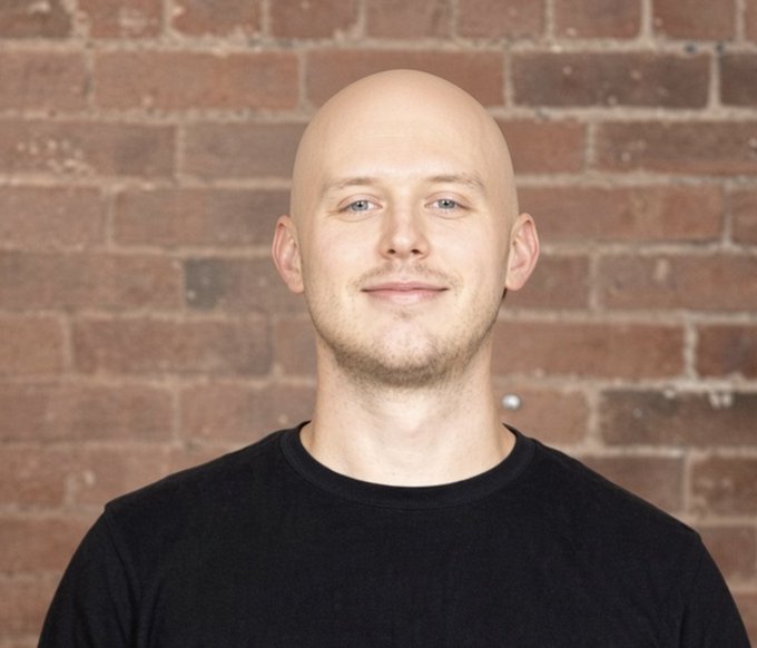 A bald man in a black t-shirt smiles softly while standing in front of an exposed brick wall, appearing to be a casual portrait or headshot. The image does not depict or relate to Brian Armstrong, nor does it contain any visible text, names, or branding that would connect it to Coinbase or the "Armstrong Trifecta" mentioned in the post.
