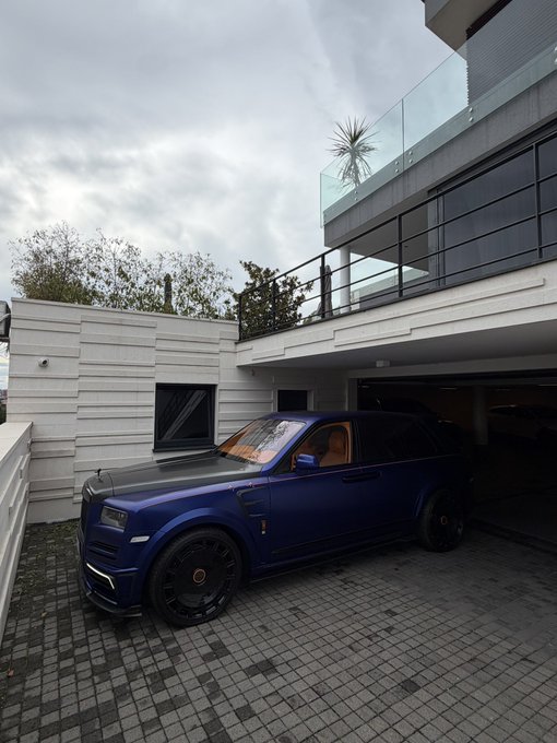 A matte blue Rolls-Royce Cullinan is parked in the driveway of a modern luxury home, exemplifying the post's message about converting cryptocurrency gains into tangible hard assets like high-end real estate and exotic vehicles. The matte blue Rolls-Royce Cullinan parked at a contemporary luxury residence visually reinforces the caption's advice to convert crypto profits into physical, appreciating assets rather than keeping wealth as numbers on a screen.
