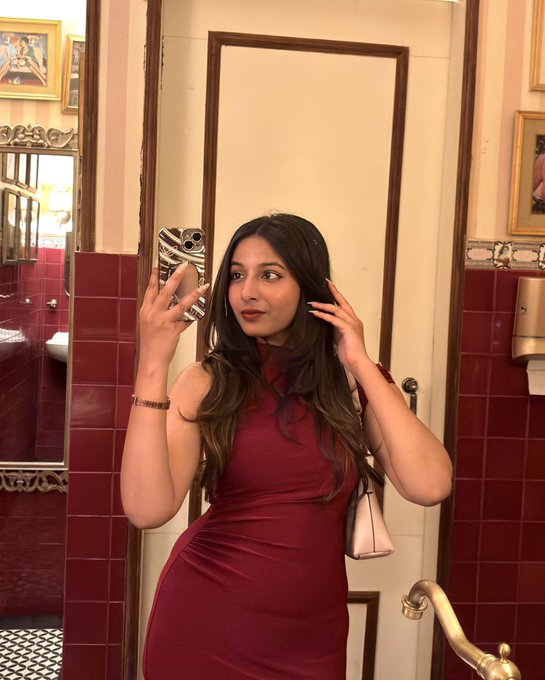 A woman in a red sleeveless dress taking a mirror selfie with her phone, standing in an ornately decorated bathroom with red tiled walls.