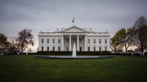 The White House is shown from the front lawn on an overcast day, with a fountain running in the foreground and trees framing the neoclassical building.