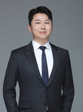 A young East Asian man in a dark suit, white dress shirt, and dark tie poses for a professional portrait against a plain gray background, smiling slightly at the camera. The polished headshot reflects the post's message of sharing an updated profile picture.
