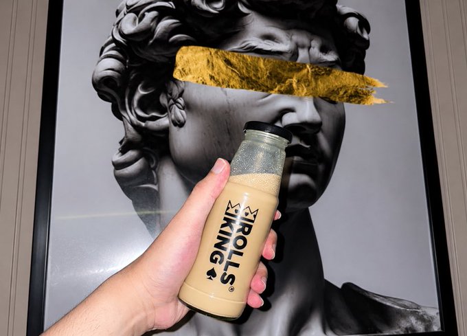 A hand holds up a bottle of "TROLLS KING" branded bubble tea in front of a framed black-and-white poster of Michelangelo's David, whose eyes are covered with a gold leaf blindfold—artfully playing on the "Trolls" theme for a cheerful "Good morning CT" post.