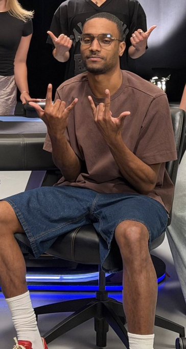 A man with glasses wearing a brown t-shirt and denim shorts sitting in a gaming chair, making hand gestures, with another person visible behind him also making hand signs.