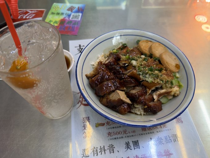 A bowl of braised pork rice with greens and fish balls sits on a placemat with Chinese text, next to a glass of iced water with a red straw on a metal table.