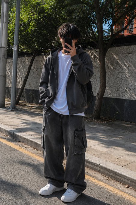 A person taking a mirror selfie on a street wearing an oversized dark gray hoodie, white t-shirt, baggy dark gray cargo pants, and white sneakers.
