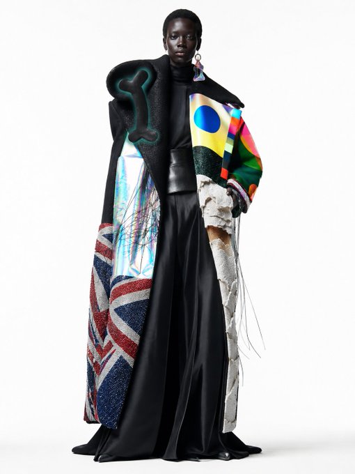 The image features a woman wearing an eye-catching outfit that includes a long, flowing dress with a unique design of red, white, blue, yellow, green, and pink colors. The dress has a high collar and a skirt made from a shiny material, giving it a distinctive appearance. She is also wearing a large coat or cape, which adds to the overall visual impact of her outfit. The woman stands out in the image due to her striking attire, making it an interesting and engaging piece for viewers who are interested in fashion and style.
