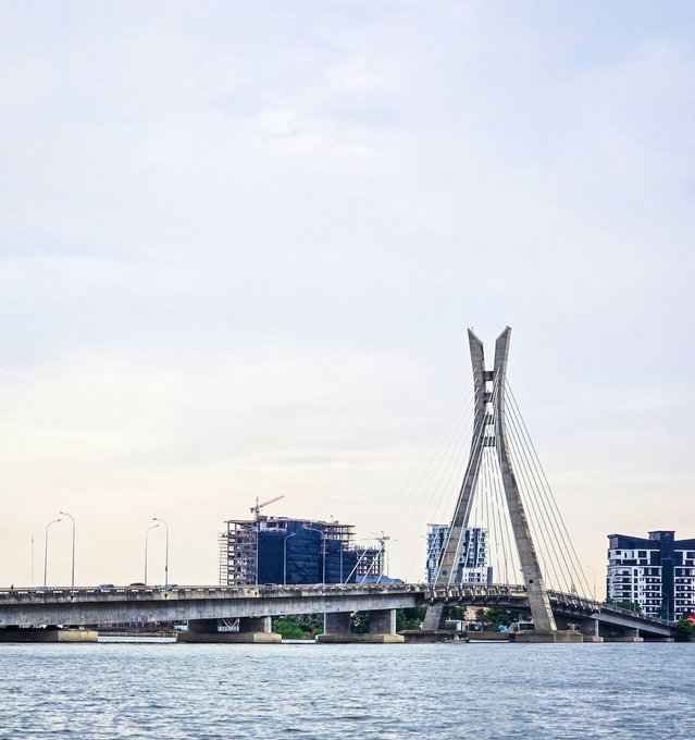 A scenic view of a modern cable-stayed bridge spanning a body of water, with city buildings and construction cranes visible in the background under a hazy sky.