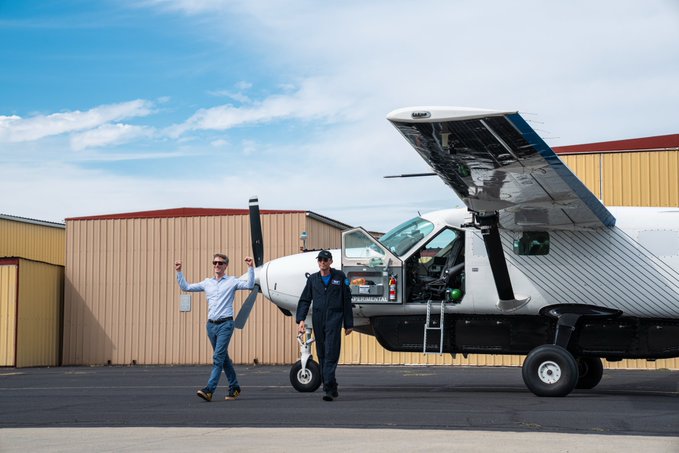 Two men walk in front of a small white propeller airplane on an airfield next to tan-colored hangars; one man has his arms raised in celebration.