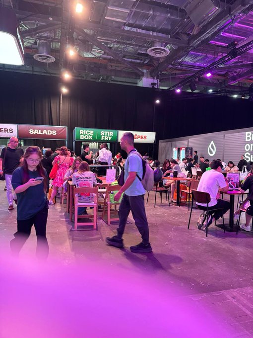 An indoor event space with food vendors like 'SALADS,' 'RICE BOX,' 'STIR FRY,' and 'CREPES,' where attendees are walking around and sitting at tables under purple and warm lighting.