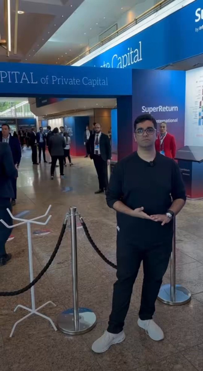 The image features a man standing next to a metal barrier with a sign that says "Super Return" above him. The man appears to be posing for a photo, possibly at an event or gathering related to the Super Return pipeline project. The background of the image shows other people present in the area as well.