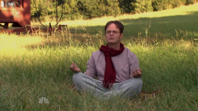 A still from the office show featuring Dwight Schrute (Rainn Wilson) meditating in a grassy field with his eyes closed and hands in a mudra position, wearing a purple shirt and red scarf.