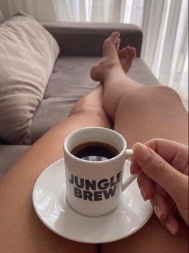 A person relaxes on a couch holding a white espresso cup filled with coffee, which has 'JUNGLE BREW' printed on it.