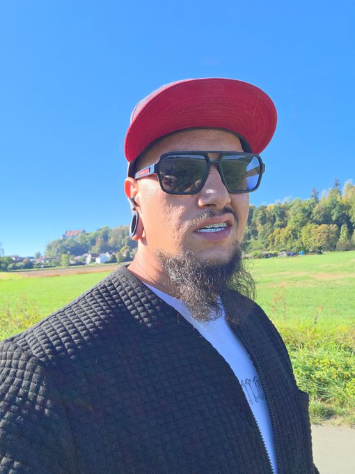 A man with a beard, wearing a red cap, black sunglasses, and a black quilted jacket, takes a selfie outdoors with a green field and hills in the background.