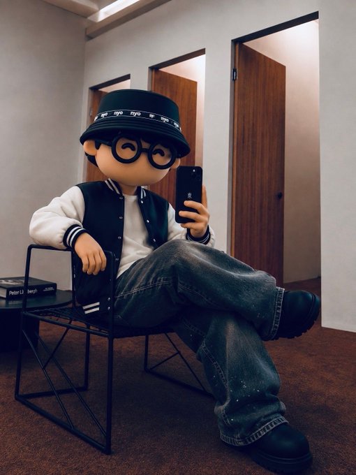 A large mascot figure resembling a cartoon character with black round glasses and a black bucket hat sits on a wire chair, holding a smartphone. Books and a wooden door are visible in the background.