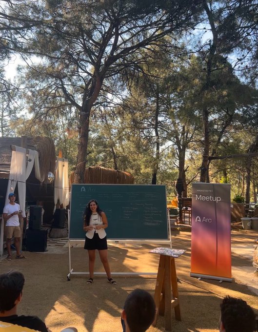 A woman stands in front of a chalkboard branded with the "Arc" logo, presenting at an outdoor event featuring a "LABORATORY Meetup" banner. The image captures her presentation at Rlay Blockchain Week 2026, where she discusses Arc's ecosystem vision alongside Circle, with attendees gathered in a forested outdoor venue.