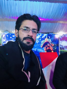 A selfie of a man with glasses and a beard wearing a black jacket, smiling at an indoor event with blue lighting, Christmas decorations, and another attendee in a Santa hat in the background.