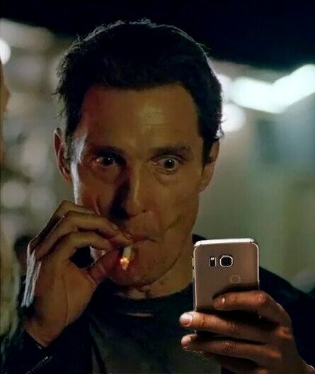 A close-up photograph of a man in low light, looking intently at a smartphone while smoking a cigarette, with blurred lights in the background.