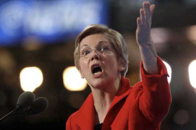 A woman in a red blazer and glasses speaks passionately at a podium, raising her arm while delivering an address. The image, appearing to show Senator Elizabeth Warren, accompanies a post criticizing the banking industry's role in money laundering to advocate for the Clarity Act.