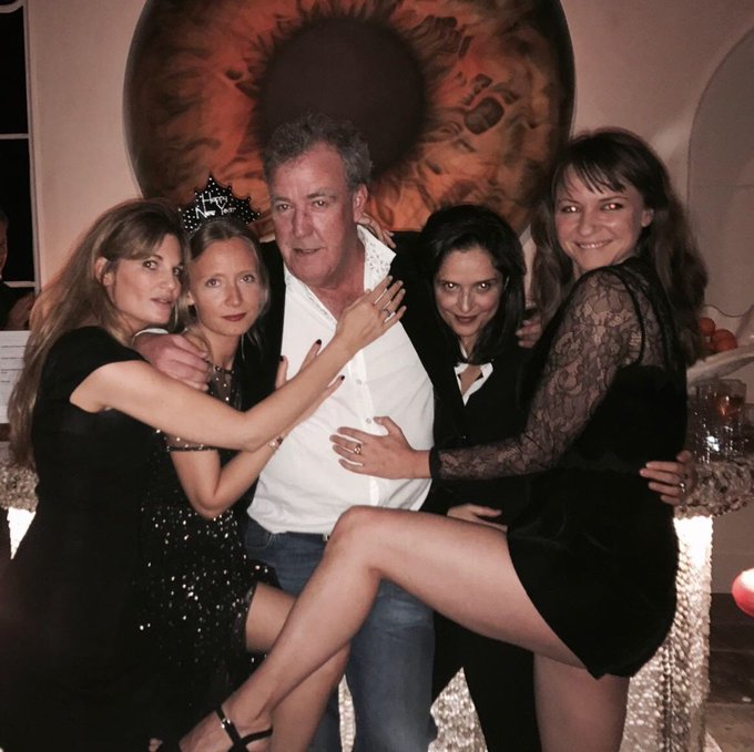 A man with gray hair, wearing a white shirt and denim jeans, stands smiling as he holds four women who are dressed in elegant black outfits. They pose together closely in front of a large decorative eye painting on the wall.