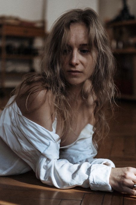 A moody portrait photograph of a contemplative woman with long, wavy dirty-blonde hair, wearing an unbuttoned white shirt draped off her shoulders as she leans on a wooden floor in a softly lit interior.