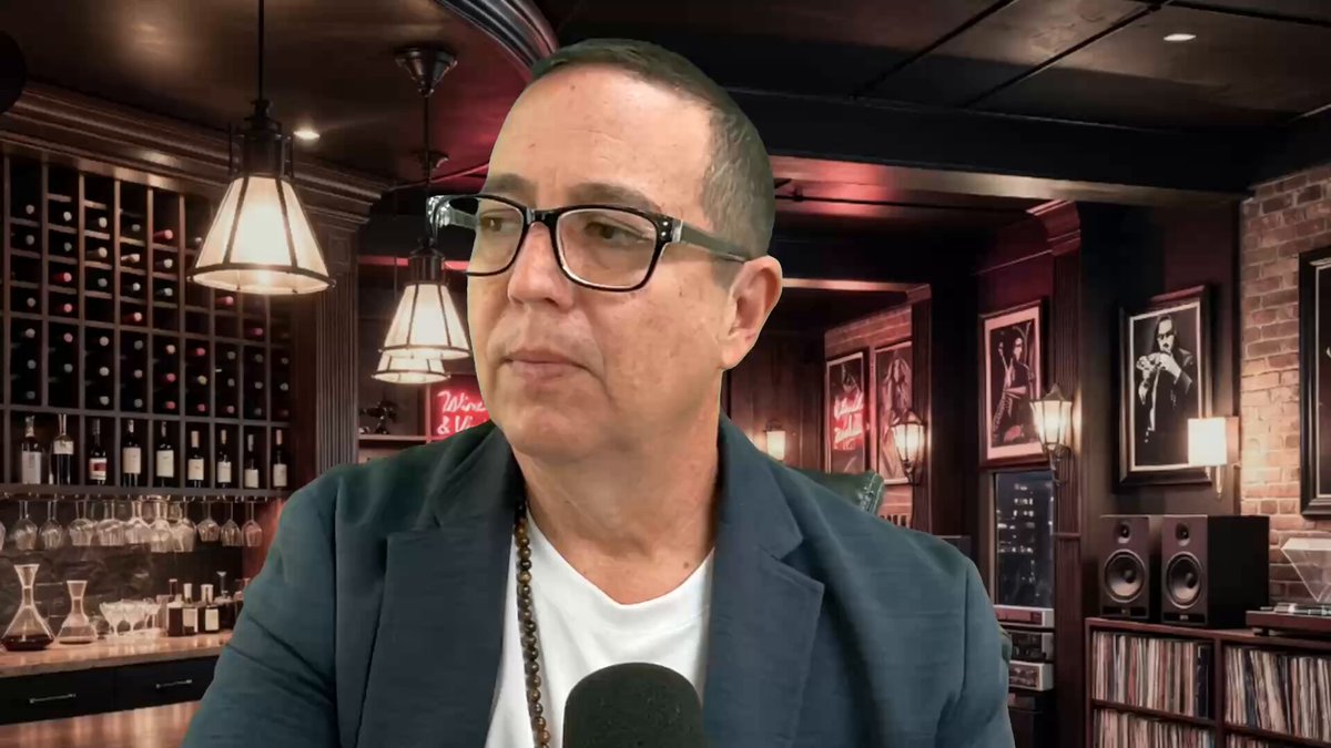 In the image, a man wearing glasses is sitting at a bar with a microphone in front of him. He appears to be talking into the microphone or recording something. The background features a wine cellar filled with numerous bottles, suggesting that this might be a wine tasting event or a bar where people gather for socializing and enjoying drinks. There are also several books visible on shelves nearby, indicating that the establishment may have an interest in literature or hosts events related to it.