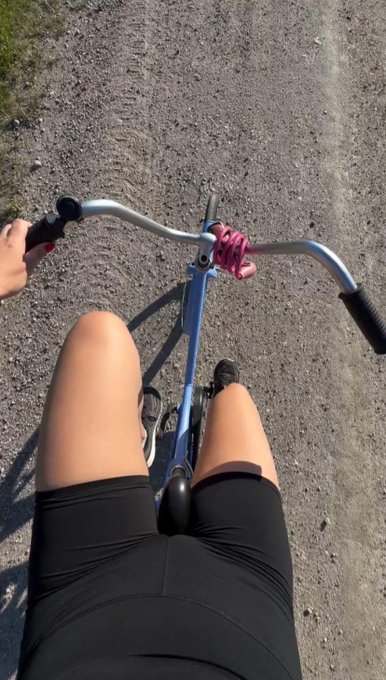 A first-person POV shot from a bicycle, showing the rider's legs in black shorts gripping the handlebars while riding down a paved road.