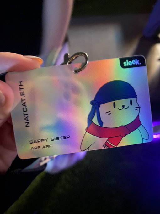 A hand holding a holographic 'Sleek' card labeled 'NATCAT.ETH' featuring a cartoon cat character called 'SAPPY SISTER' with the text 'ARF ARF'.