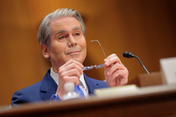 A man in a blue suit holds up a pair of glasses while smiling in what appears to be a hearing room with a microphone visible.
