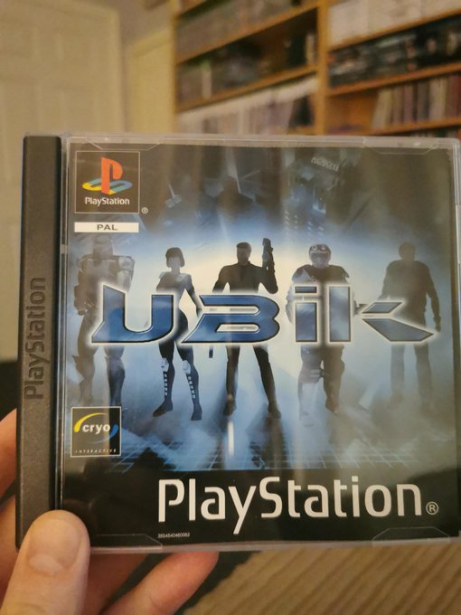 A hand holds a PlayStation game case for "UBIK," a PAL region title published by Cryo Interactive, featuring silhouettes of characters on the cover. The user is humorously questioning what the title means, playing on "UBIK" being a Philip K. Dick novel that is famously confusing and ambiguous.