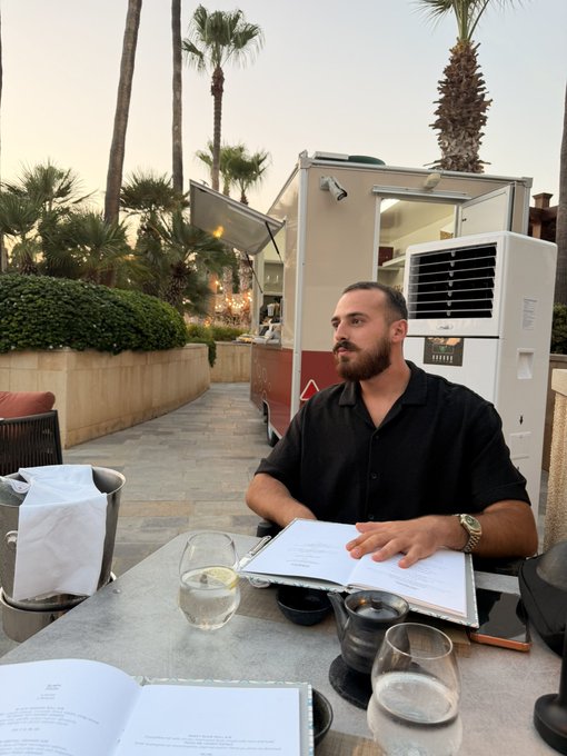 The image shows a bearded man in a black shirt seated at an outdoor restaurant table reviewing menus, with a vintage food truck and palm trees in the background. A wine glass, water glass, and what appears to be a tablet rest on the table in front of him, suggesting a sophisticated dining setting during golden hour.