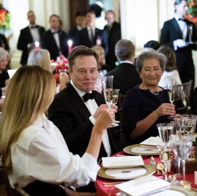 Elon Musk in a tuxedo sits at a formal dinner, toasting with a champagne glass alongside other guests.