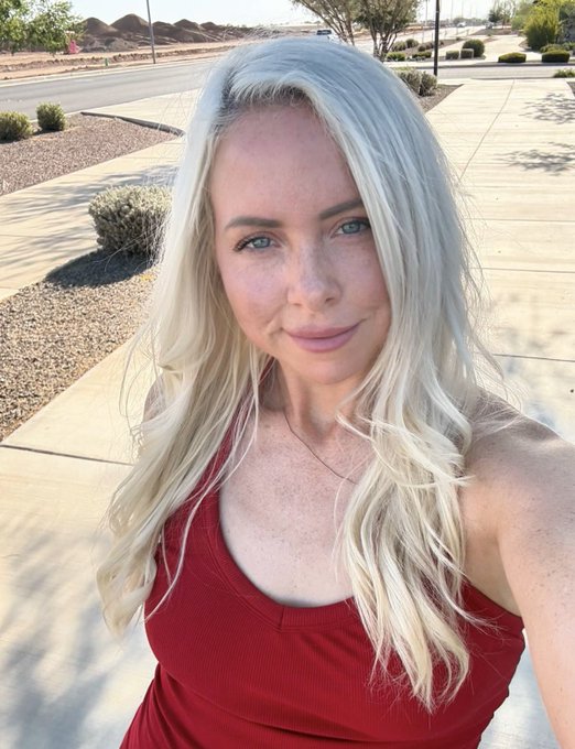 A selfie of a blonde woman wearing a red tank top, taken outdoors on a sidewalk in a desert-like neighborhood with sparse landscaping.