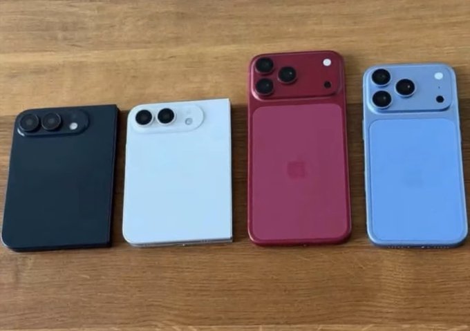 Four smartphones displayed face-down on a wooden surface, showing what appear to be iPhone models in different colors including black, white, red, and blue, with varying camera module designs.