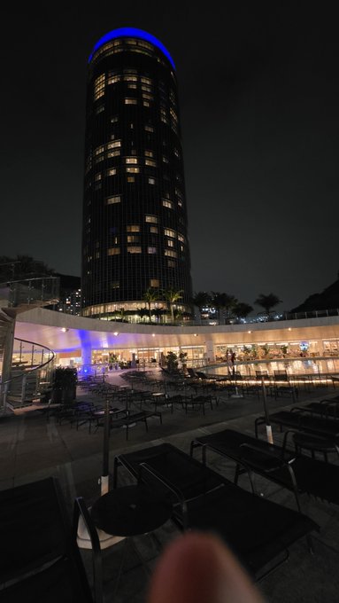 A nighttime photograph of a tall cylindrical skyscraper illuminated with blue lights at the top, overlooking a multi-level outdoor plaza with empty tables and chairs.