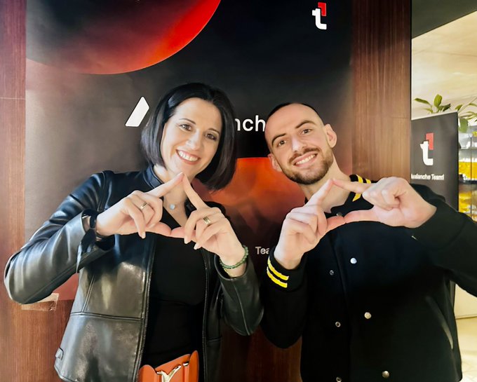 A man and woman smiling and posing together indoors in front of a backdrop featuring logos, with the woman in a black leather jacket and the man making a heart shape with his hands.