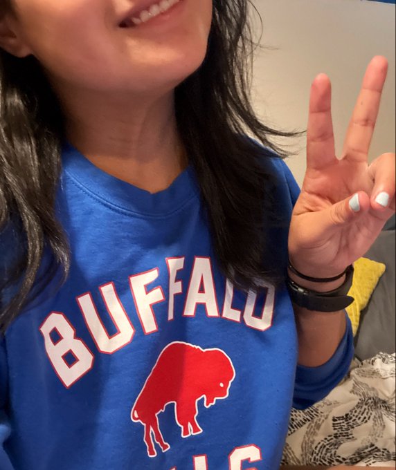 A smiling person wearing a blue Buffalo Bills t-shirt with a red buffalo logo, making a peace sign with their right hand.