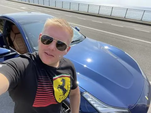 A man wearing sunglasses and a black Ferrari t-shirt takes a selfie in front of a blue luxury car on a bridge, though the image itself doesn't visually relate to the post about Suszk's disappearance or the BitBay-to-ZondaCrypto rebranding.