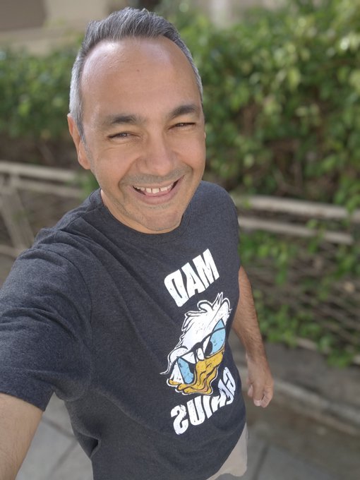 A man smiles broadly while taking a selfie, wearing a dark t-shirt featuring a duck graphic and the text 'DAM' and 'GUARD'.