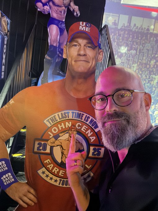 A bald man with glasses and a beard takes a selfie next to a life-sized cardboard cutout of wrestler John Cena wearing an orange shirt that says 'THE LAST TIME IS NOW JOHN CENA FAREWELL TOUR 20 25' and a red 'U CAN'T ME' cap.