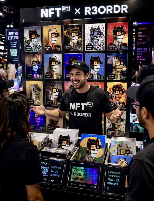 A smiling man wearing a black 'NFT NYC x R3ORDR' t-shirt and cap stands at a merchandise booth displaying pixel-art framed prints and album covers, advertising 'LIMITED EDITION' items with a QR code.