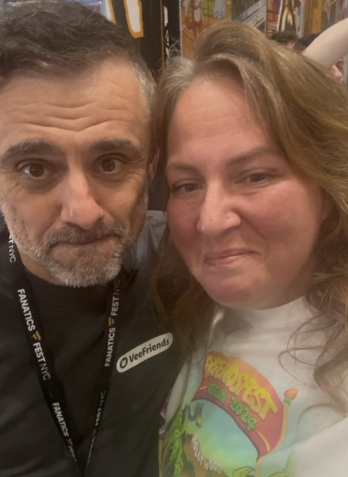 A selfie of a man and a woman at what appears to be a Fanatics Fest NYC event, with both wearing event lanyards; the woman's shirt features 'Fanatics Fest 2024' branding.