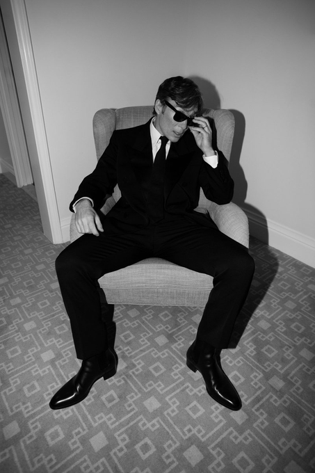 A black and white photo of a man in a suit and sunglasses sitting in a chair, adjusting his glasses while smiling.