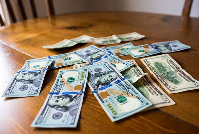 A scattering of US currency, predominantly $50 and $100 bills bound by blue currency straps, lies spread across a wooden table surface.