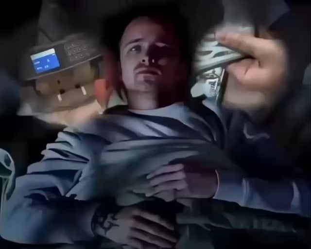 A man in a light blue sweatshirt appearing to be in a vehicle, holding what looks like money or papers, with a distorted, low-quality image effect applied.