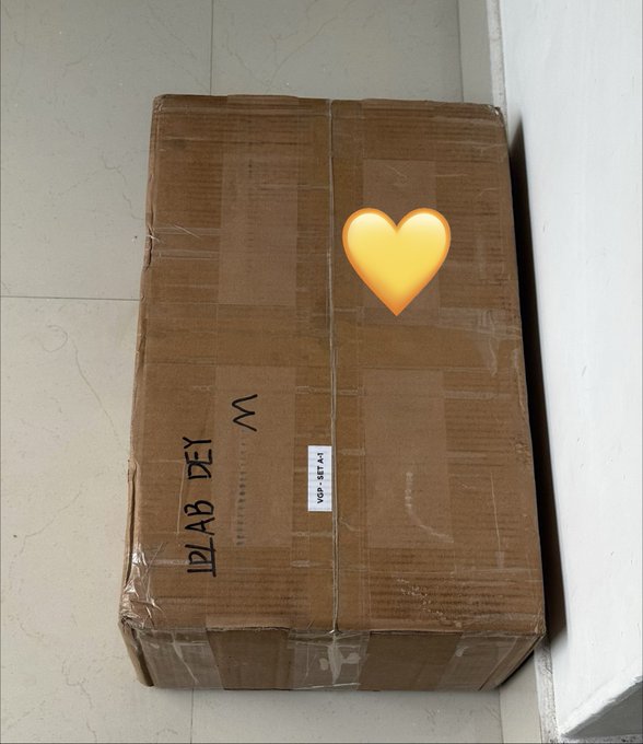 Photograph of a large sealed cardboard box on a tiled floor with a heart emoji overlay and text reading 'M' and 'X12L B71'.