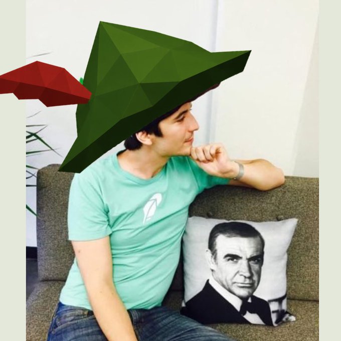 A man sits on a couch next to a pillow featuring Sean Connery's face, wearing a low-poly 3D-printed Yoda hat and holding a red lightsaber.