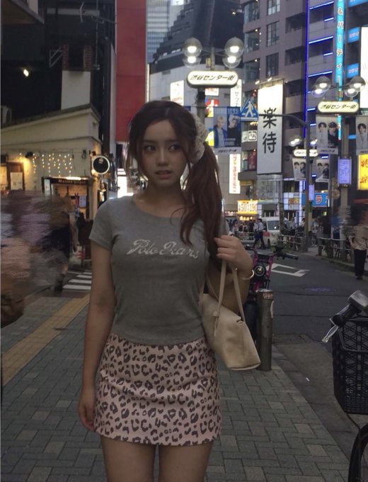 A young woman wearing a gray 'Pilots in Paris' t-shirt and a leopard print skirt stands on a city street at night, holding a beige tote bag.